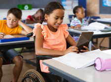 In einem Klassenzimmer sitzen mehrere Kinder an ihren Tischen und arbeiten konzentriert. Im Vordergrund sitzt ein Mädchen im Rollstuhl, das ein Tablet in den Händen hält und lächelt. Auf ihrem Tisch liegen Schulmaterialien wie Hefte und ein Federmäppchen.