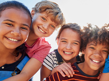 Eine Gruppe von sechs fröhlichen Kindern steht dicht beieinander und lächelt in die Kamera. Sie sind bunt gekleidet.