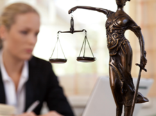 Das Bild zeigt eine Frau in einem Büro, die an einem Laptop arbeitet. Im Vordergrund steht eine Statue der Justitia, die Göttin der Gerechtigkeit, mit einer Waage in der einen und einem Schwert in der anderen Hand. Die Frau im Hintergrund ist unscharf dargestellt, während die Statue scharf fokussiert ist. Auf dem Schreibtisch vor ihr liegt ein aufgeschlagenes Buch.