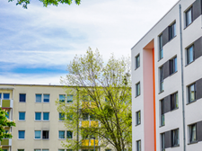 Das Bild zeigt eine städtische Wohngegend mit modernen Mehrfamilienhäusern. Die Gebäude haben eine helle Fassade, wobei eines der Häuser durch orangefarbene Akzente hervorsticht. Die Fenster sind gleichmäßig angeordnet und einige Balkone sind zu sehen.